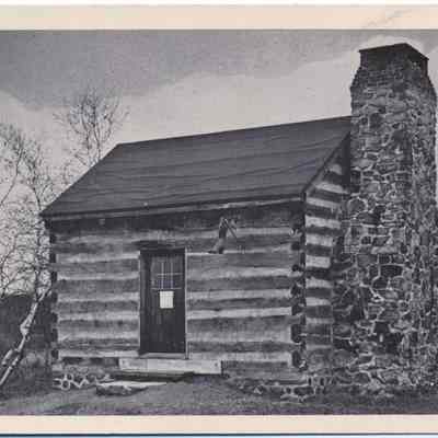 Shoemaker Shop at Old Museum Village Of Smith's Clove: This monochrome photo postcard shows an old log cabin with a prominent stone chimney on its right side, positioned in a tranquil rural setting under a cloudy sky. The cabin's rustic appearance, featuring exposed log walls and a simple black door with a window, portrays a historical architecture typical of early American or pioneer times. The bare trees in the background and a sparse landscape enhance the old-fashioned feel, suggesting the photo could be from an earlier century, capturing a piece of history preserved at Old Museum Village Of Smith's Clove.