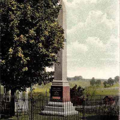 “Hambletonian's Monument, Chester, N. Y.” Front: The image depicts the Hambletonian Monument located in Chester, New York. This historic structure, prominently featured in a lush and scenic park setting, includes a tall, obelisk-like stone pillar standing on a decorative base that is inscribed and partly shaded by a vibrant green tree. The monument is encircled by a well-maintained iron fence that enhances the reverence of the site. The background shows an expansive grassy field stretching towards a line of trees under a partly cloudy sky, emphasizing the peaceful and pastoral ambiance of the location. The overall appearance suggests this might be a commemorative site dedicated to an important figure or event in the region's history.