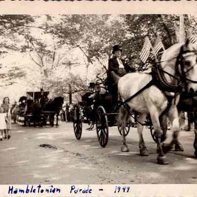 Hambletonian Parade - 1949: The black and white image depicts the Hambletonian Parade, showcasing a scene with people lined along a shaded street watching the parade. The focus is on Frank Scriver wearing a top hat driving a horse-drawn carriage, pulled by two white horses adorned with harnesses and flags. Spectators of various ages, including children and adults, observe the procession with visible interest.