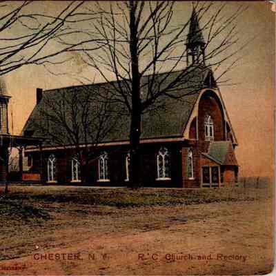 “Chester, N.Y. R.C. Church and Rectory” Front. CHS-019957-4: The image captures a serene historic scene in Chester, New York, featuring two prominent buildings: a picturesque Roman Catholic Church and its adjacent rectory. On the left, the rectory is a large, multi-story house with a welcoming front porch and distinct yellow siding, contrasting with its darker, decorative trimmings. To its right stands the church, characterized by its classic Gothic architecture, including pointed arch windows and a steep, pointed roof with a bell tower prominently rising at its front. The church's brick facade pairs harmoniously with its tranquil rural surroundings, where leafless trees subtly frame the buildings, hinting at a late autumn or winter season. This charming early 20th-century postcard, marked as "Made in Germany," provides a nostalgic glimpse into the architectural and community heritage of the time.