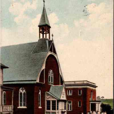“Catholic Church, Chester. N. Y.” Front. CHS-019957-1: The image depicts the Catholic Church in Chester, NY, as indicated by the text at the top. This historical postcard shows a charming, old-fashioned church with a steeply pitched roof and a prominent steeple. The architecture features gothic-style windows and a bright red facade, supplemented by white trimmings around the windows and doors, which contribute to its striking appearance. Adjacent to the church, a larger brick building can be seen, which possibly serves as a rectory or parish hall. A small group of people is gathered at the entrance of this annex, hinting at some community activity or gathering. The sky above is scattered with clouds, suggesting a serene day. The lawn in front of the church is well-manicured, lined with a neat pathway leading to the buildings, enhancing the welcoming and peaceful ambiance of the religious complex.
