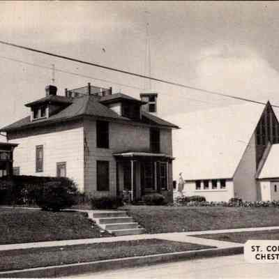“St. Columba Church Chester, New York” Front. CHS-019957-3: The image captures a scene in Chester, New York, featuring St. Columba Church, distinguished by its sharp, triangular roof and clean, modern architectural lines. Next to the church, a tastefully designed statue accentuates its religious significance. In the foreground, a large traditional house, possibly a parsonage, exhibits classic features with multiple stories, a wrap-around porch, and a prominent tower-like structure. Utility lines cross the sky above, and well-maintained lawns complement the urban landscape, lending an air of calm and order. The monochrome tone of the photograph provides a timeless quality, reflecting the historical context of the area.