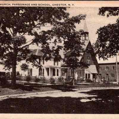 “St. Columbia Church, Parsonage and School, Chester, N.Y.” Front. CHS-019957-2: The image depicts a serene view of St. Columbia Church, along with its parsonage and school, located in Chester, New York. In the foreground, a shaded street is visible, lined with mature trees that partially obscure and dapple the sunlight on the structures. On the left, a traditional, white parsonage house, with its prominent porch, can be seen neighboring a larger building presumably used for educational purposes. On the right, the church is more distinctly Gothic in architecture, featuring a pointed arch window and a steeply pitched roof topped with a slender spire that punctuates the skyline. These buildings, representative of community and architecture from a bygone era, are nestled in a tranquil setting that conveys a sense of historical and spiritual significance. The photo, monochromatic and slightly grainy, adds to the nostalgic essence of the scene.