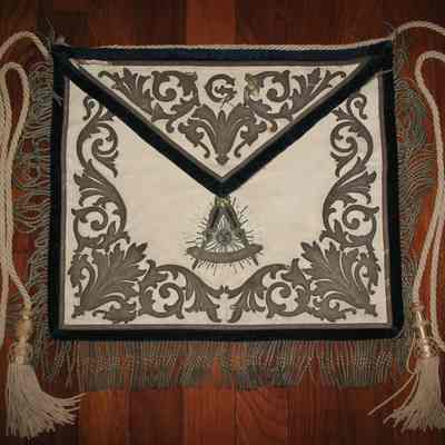 Apron: District Lecturers apron. White leather, brocade Past Master's emblem and other designs. Blue velvet border, tassels same color brocade. White cord. Under flap: "Presented by the 24th District to Bro. John W. Moore District Lecturer". Additional information in Source Binder.