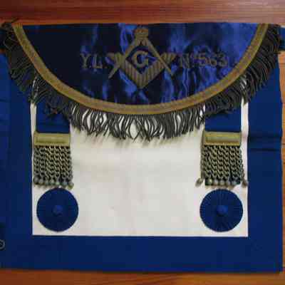Apron: Made in 1923 by Thomas McIntyre of Dumbarton, Scotland. Given to Jean R. Pritcherd for her husband James H. Pritcherd. 14" x 15 1/2" white leather. In brocade "Y.L.-Square & CompassNo563" on the flap. Blue satin lining. Blue satin B=borders, adjustable belt.