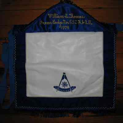 Apron: Past Masters Apron, machine embroidered: Past Masters emblem on flap, Ornan, P.M. - under flap, William G. Thomas Ornan Lodge No. 652, 1978. 15" x 13". White with blue velvet border and 1 1/2" fringe on the bottom of flap and bottom of body.