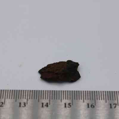 Closeup of basaltic glass cortex flake with ruler: The image shows a small basaltic glass cortex flake resting on a white surface, next to a ruler for scale.