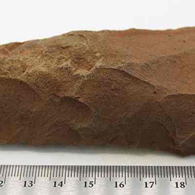 Closeup profile view of adze blank with ruler: The image shows a roughly hewn, reddish-brown stone tool blank, displayed against a neutral background with a ruler measuring its length, indicating it is approximately 12 cm.