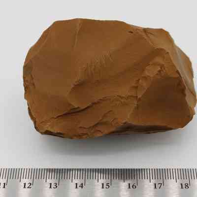 Close up of adze preform with ruler: A rough, brown stone sits on a flat surface, with a metal ruler beneath it, suggesting a tool-making context likely related to shaping.