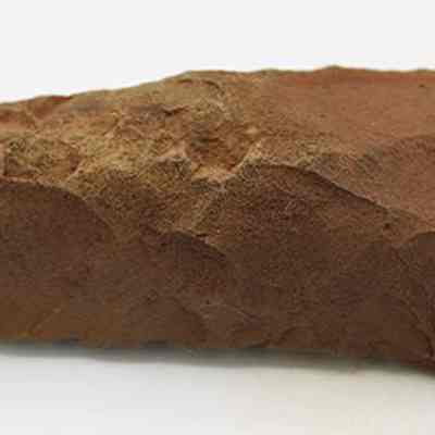 Profile of adze blank: The image shows a reddish-brown, angular stone tool with rough edges and a pointed tip, sitting on a plain white background, suggesting an ancient artifact.