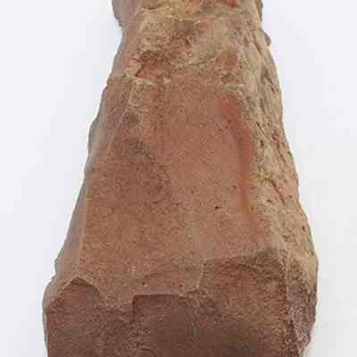 Closeup lengthwise view of adze blank: The image depicts a rough, reddish-brown stone with sharp, uneven edges, likely shaped by  humans for cutting or scraping purposes.