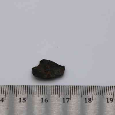 Tiny basaltic glass flake with ruler: A small basaltic glass flake rests next to a centimeter ruler, illustrating its size and showcasing its dark, rugged surface.