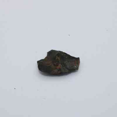 Closeup of basaltic glass flake: The image shows a small, irregularly shaped basaltic glass flake resting on a plain, light-colored surface.