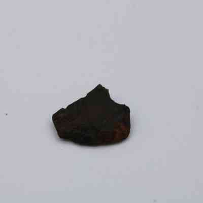 Closeup of tiny basaltic glass flake: A small, dark fragment of basaltic glass flake lies centered on a plain, light background, highlighting its rugged texture and irregular shape.