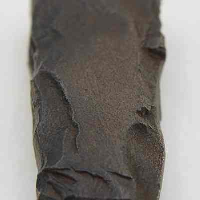 Close up lengthwise view of an adze fragment: The image shows a roughly rectangular, dark stone tool fragment with jagged edges,, sitting on a light, plain background.