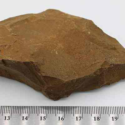 Closeup of basalt drill and ruler: The image shows a brown, roughly triangular stone drill, placed on a flat surface, beside a metal ruler for scale, indicating its size.