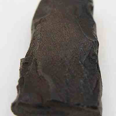 Closeup lengthwise view of adze fragment: The image showcases a close-up of a dark, rectangular-shaped stone with a rough texture, highlighting its uneven surface details and natural imperfections on a pale background.