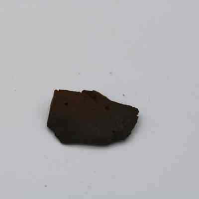 Closeup of basaltic glass flake: A small, dark basaltic glass flake lies isolated against a plain white background, displaying an irregular shape and rough surface texture.