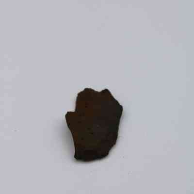 Closeup of basaltic glass flake: A jagged, dark basaltic glass flake rests against a light background, showcasing a rough, uneven texture with small surface pits.