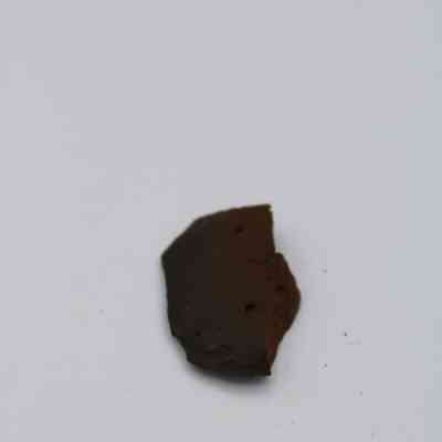 Closeup of basaltic glass flake: A small, dark brown basaltic glass flake is resting against a plain, white background, highlighting its rough texture.
