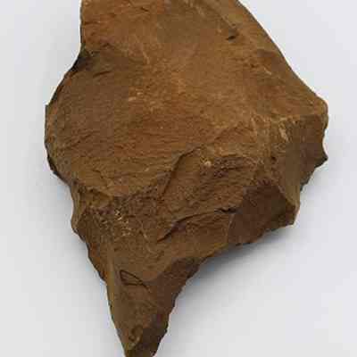 Lengthwise view of basalt drill with ruler: The image shows a brown, irregularly shaped stone drill with rough, uneven surfaces, placed next to a ruler for scale under bright lighting, highlighting its texture.