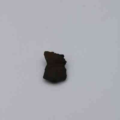 Closeup of basaltic glass flake: The image shows a small, dark basaltic glass flake with irregular edges on a plain, light background.