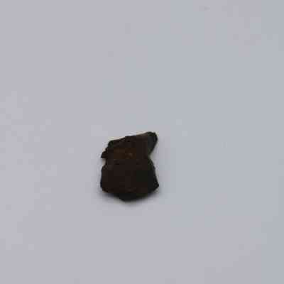 Closeup of basaltic glass flake: The image shows a small, dark piece of basaltic glass flake lying on a plain white background, highlighting its rough texture and irregular shape.