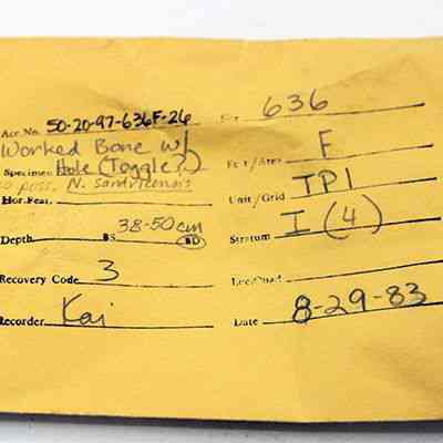 Artifact envelope for possible bone toggle: The image shows a yellow envelope with handwritten text, detailing an archaeological record of a worked bone specimen.