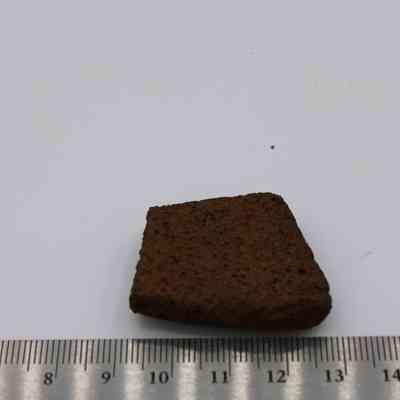 Closeup of file fragment and ruler: The image shows a small, textured fragment of basalt rock placed on a white surface, alongside a metallic ruler for scale.