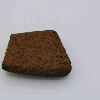 Closeup of basaltic file fragment: The image shows a porous, rectangular fragment of basalt rock, characterized by a rough, textured surface, sitting on a plain, light-colored background.