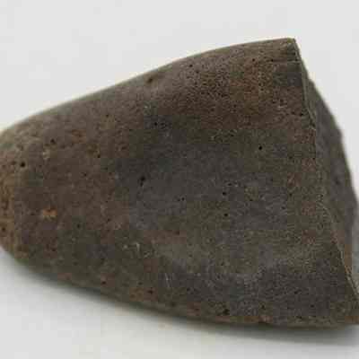 Closeup side profile view of a hammerstone: The image shows a dark, weathered stone with a roughly conical shape, lying on a white surface.