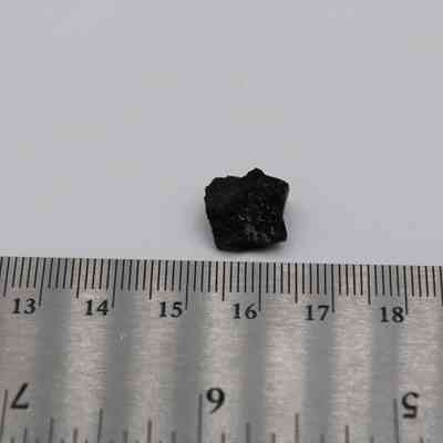 Closeup of basaltic glass flake with ruler: A small basaltic glass flake is positioned above a metallic ruler, showing its size and texture against a plain white background.