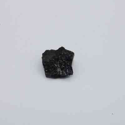 Closeup of basaltic glass flake: A small fragment of basaltic glass flake rests on a plain white surface, showcasing its dark, slightly textured and uneven surface.
