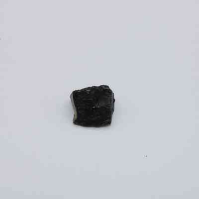 Closeup of basaltic glass flake: A small, dark basaltic glass flake rests on a light, neutral background, showcasing its rough texture and subtle shine.