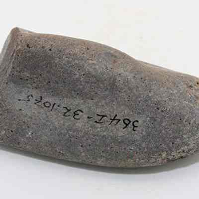 Closeup side profile of hammerstone with visible alternate id number: The image shows a smooth, tapered stone artifact, possibly an ancient tool or implement, placed on a plain surface with an identification number inscribed on it in black ink.