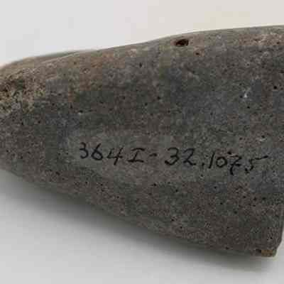 Closeup of hammerstone with visible alternate id number: The image shows a cone-shaped gray stone with a rough texture lying on a white surface. A series of numbers, 364I-32.1075, is handwritten in black ink.