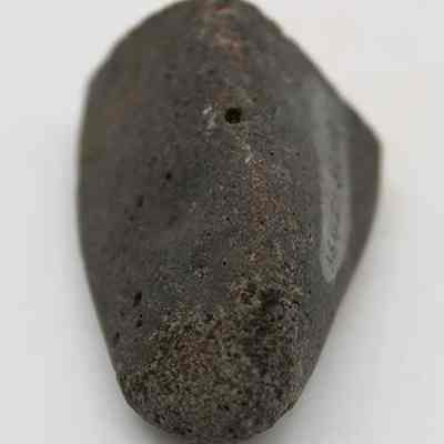 Closeup lengthwise view of a hammerstone: The image shows a rough, dark gray stone with an irregular shape, likely a piece of basalt, lying against a light background.