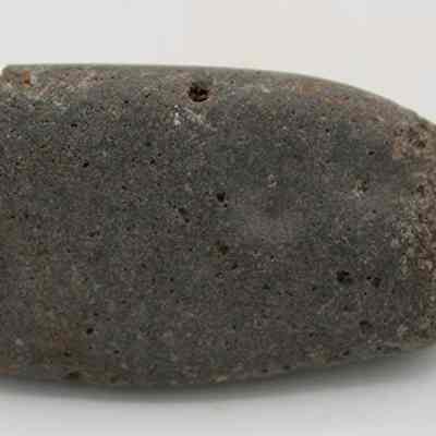 Closeup side profile of a hammerstone: The image shows a weathered, dark gray stone with a rounded, tapering shape and a rough, textured surface, likely an ancient tool or artifact such as a hammerstone.