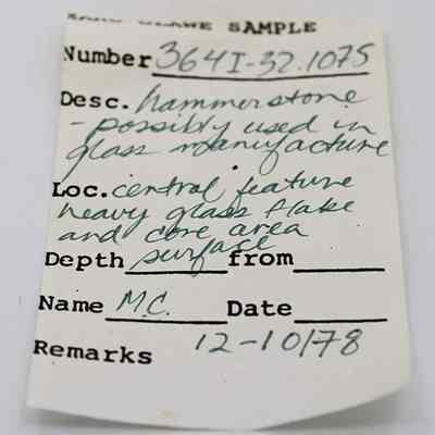 Artifact slip for hammerstone: The image shows a handwritten label listing details of an artifact. It includes a sample number, description of a hammerstone potentially for glass manufacture.