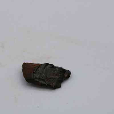 Closeup of basaltic glass flake: The image shows a small, irregularly shaped flake of basaltic glass, resting on a gray surface with minimal texture.