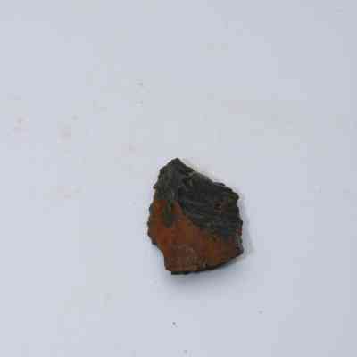 Closeup of basaltic glass flake: A small, irregularly shaped basaltic glass flake lies on a plain white surface, showcasing a dark, rugged texture.
