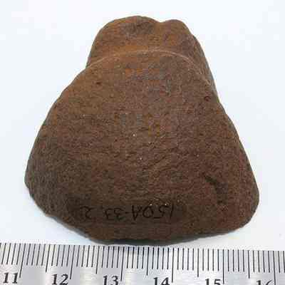 Grooved type sinker or polishing stone with ruler: The image shows a rough, brown stone fragment, possibly a sinker or polishing tool, resting on a surface with a ruler for size reference.