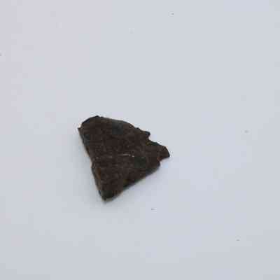 Basaltic glass flake: A small piece of baslatic glass rests on a plain white surface, showcasing its dark, rough texture and angular shape.