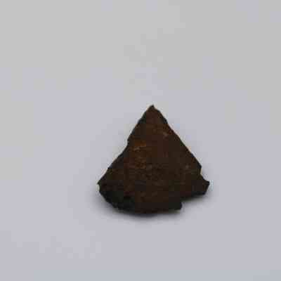 Triangular piece of basaltic glass: A triangular piece of basaltic glass, dark brown and textured, lies on a plain white surface, emphasizing its rugged and weathered appearance.