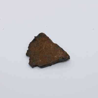 Triangular piece of basaltic glass flake: A piece of basaltic glass, with a jagged shape, rests on a plain white surface, displaying a dark, rough texture and earthy hue.