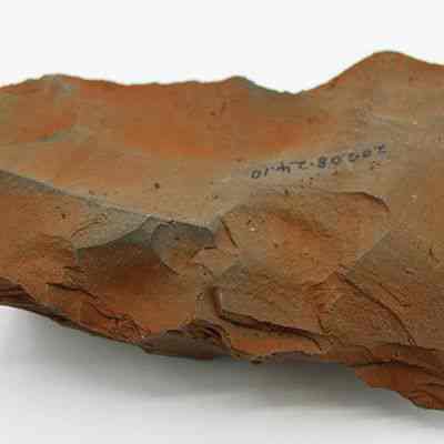 Adze blank with visible alternate id number: The image shows a reddish-brown stone tool or artifact with rough, uneven surfaces and visible markings, likely indicative of its archaeological significance.