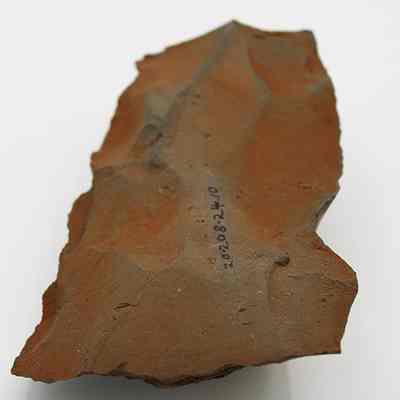 Lengthwise view of adze blank with visible alternate id number: The image shows an orange-brown triangular stone with uneven surface texture and a number sequence inscribed on it.