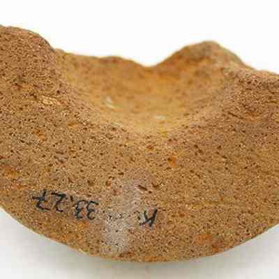Closeup of fragment of cup or mortar with alternate id number visible: The image shows a rough, arc-shaped fragment of pottery, possibly from a cup or mortar, characterized by its coarse texture and visible surface markings.
