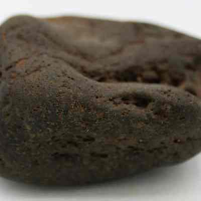 Side of hammerstone: The image shows a dark, smooth, irregularly shaped rock with a rough texture and small indentations on its surface, set against a plain white background.
