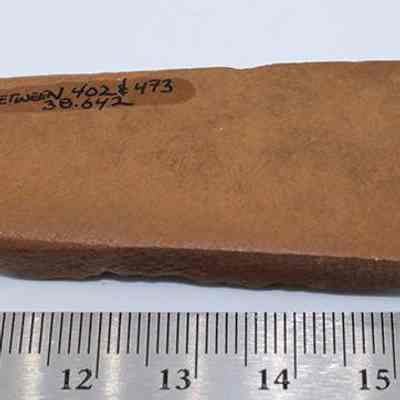 Bottom side of basalt adze and ruler: A rectangular brown stone adze, with text "BETWEEN 402 & 473 38.642" written on it, is placed above a ruler.